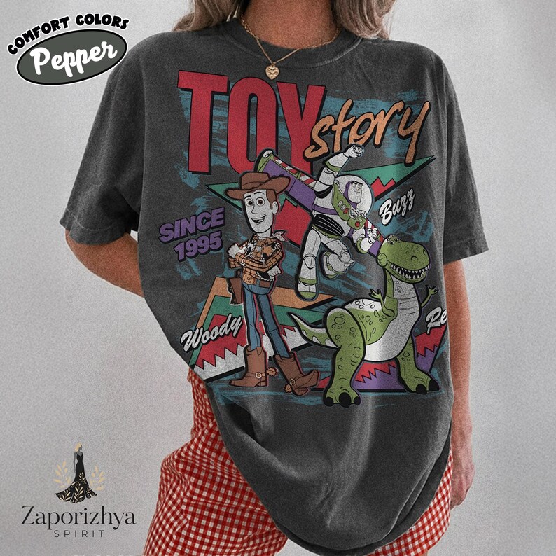 May include: A charcoal gray short-sleeve t-shirt featuring a graphic print of Toy Story characters Woody, Buzz Lightyear, and Rex. The design includes the text Toy Story, Since 1995, Woody, and Buzz, set against colorful geometric shapes.
