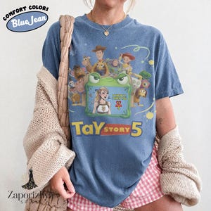 May include: A blue jean colored t-shirt featuring a graphic print of Toy Story characters surrounding a green frog frame. Inside the frame is a blonde woman with the text I KNEW IT I KNEW YOU TS STARS 5. Below the graphic is the text TAY STORY 5.
