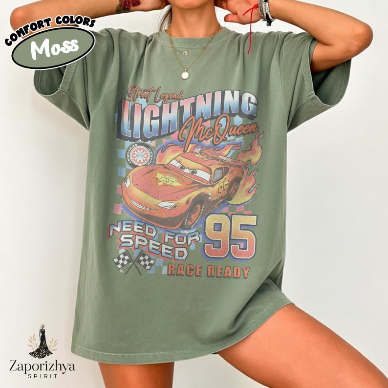 May include: Olive green t-shirt featuring a graphic of Lightning McQueen from the movie Cars. The design includes the text "Street Legends Lightning McQueen 95" and "Need for Speed Race Ready". The shirt has a relaxed fit.