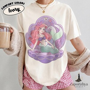 May include: An ivory t-shirt featuring a graphic illustration of a mermaid with long pink hair, a purple seashell top, and a green tail, centered within a large purple seashell frame. The design includes small pink starfish and seaweed accents.