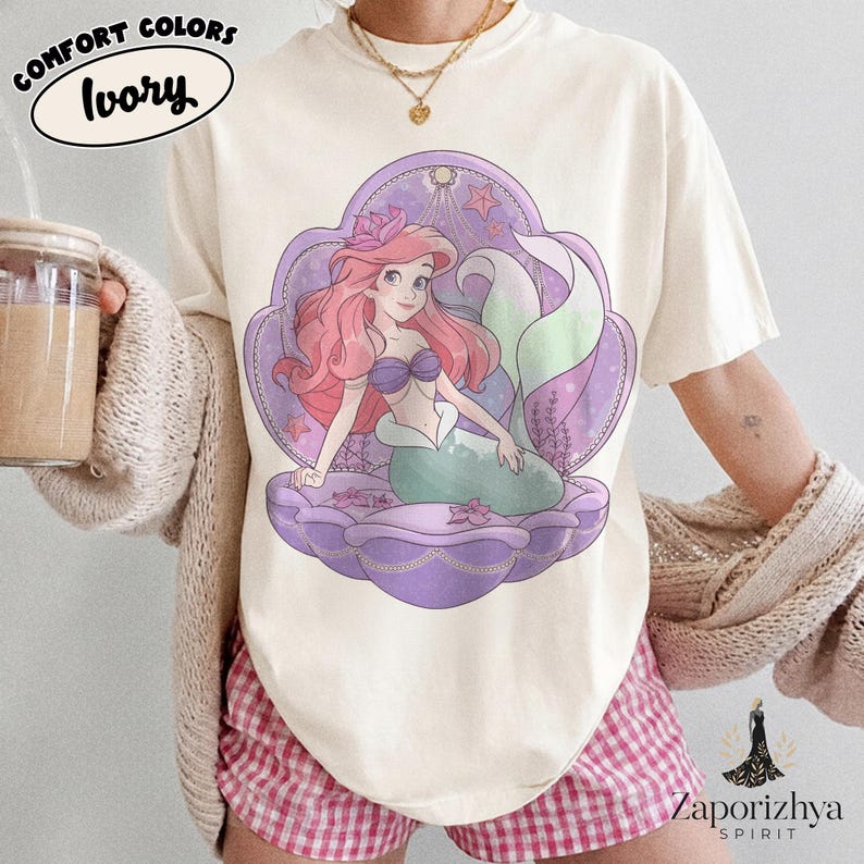 May include: An ivory t-shirt featuring a graphic illustration of a mermaid with long pink hair, a purple seashell top, and a green tail, centered within a large purple seashell frame. The design includes small pink starfish and seaweed accents.