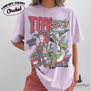 May include: A light purple short-sleeve t-shirt featuring a graphic print of Woody, Buzz Lightyear, and Rex from Toy Story. The text reads Toy Story, Since 1995, Woody, Buzz, and Rex. The shirt has a crew neckline and a relaxed, loose-fitting silhouette.
