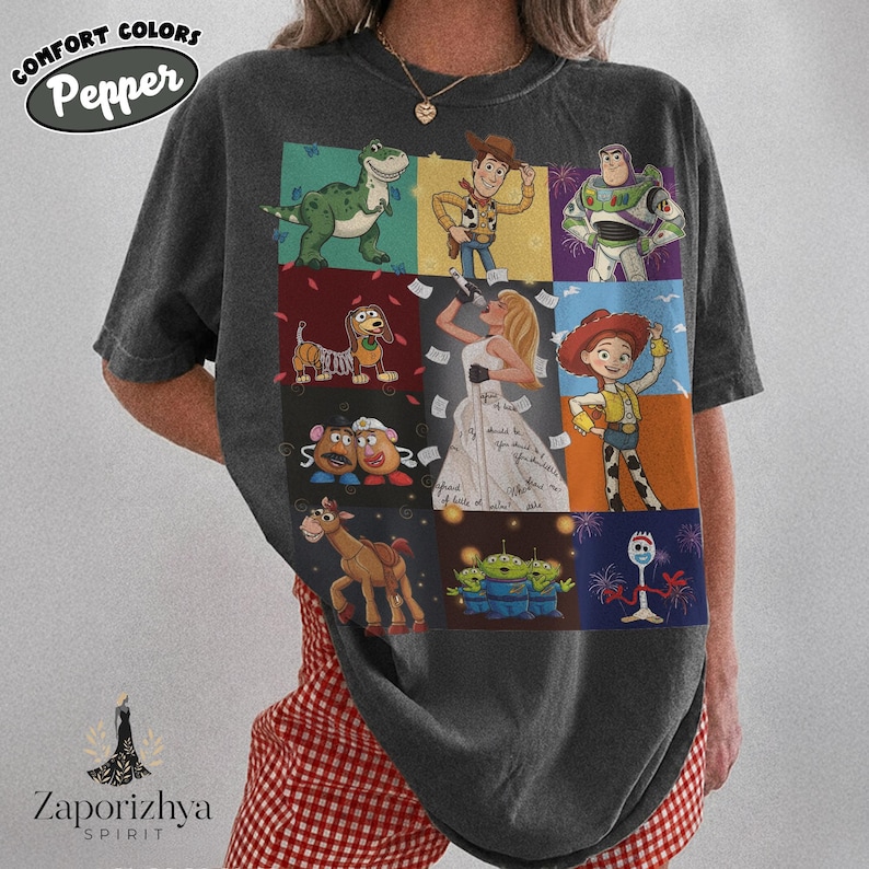 May include: A charcoal gray short-sleeve t-shirt featuring a grid of Toy Story characters including Woody, Buzz Lightyear, Jessie, Rex, Slinky Dog, Mr. and Mrs. Potato Head, Bullseye, the Aliens, Forky, and a central figure of a woman singing.