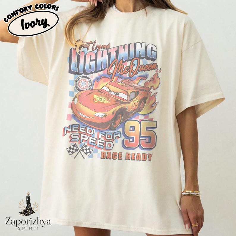 May include: Ivory-colored t-shirt featuring a graphic of Lightning McQueen from the movie Cars. The design includes the text "Great Legend Lightning McQueen", "Need for Speed", and "Race Ready" with the number 95.