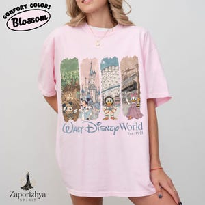 May include: A pale pink t-shirt with a graphic print of Disney characters and park icons. The design includes Mickey, Minnie, Donald, and Daisy Duck, with "Walt Disney World" and "Est. 1971".