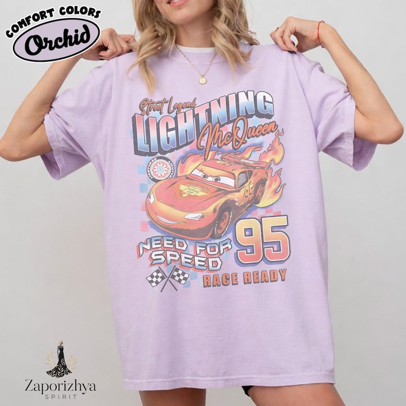 May include: A light purple t-shirt featuring a graphic of Lightning McQueen from the movie Cars. The design includes the text "Street Legend Lightning McQueen", "Need for Speed", and "Race Ready", with the number "95". The shirt has the "Comfort Colors" and "Orchid" logos.