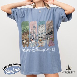 May include: Blue jean Comfort Colors t-shirt with a white collar. The front displays a colourful graphic of Disney characters and landmarks, with the text "Walt Disney World Est. 1971". The shirt has short sleeves and a relaxed fit.