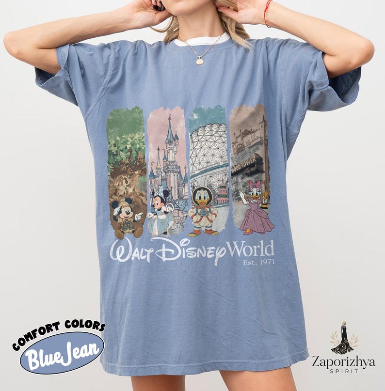 May include: Blue jean Comfort Colors t-shirt with a white collar. The front displays a colourful graphic of Disney characters and landmarks, with the text "Walt Disney World Est. 1971". The shirt has short sleeves and a relaxed fit.