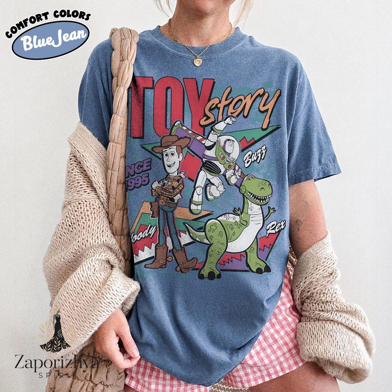 May include: A blue jean colored t-shirt featuring a graphic print of Woody, Buzz Lightyear, and Rex from Toy Story. The design includes the text Toy Story, 1995, Woody, Buzz, and Rex with geometric shapes in red, green, and purple behind the characters.