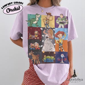 May include: A light purple short-sleeve t-shirt featuring a grid of nine panels displaying various animated characters, including a green dinosaur, a cowboy, a space ranger, a dog, a singer, a cowgirl, potato figures, a horse, aliens, and a fork character.