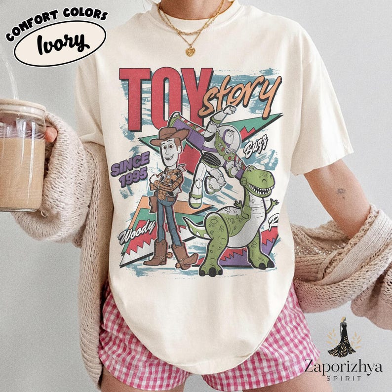 May include: An ivory short-sleeve t-shirt featuring a graphic print of the characters Woody, Buzz Lightyear, and Rex from Toy Story. The design includes the text TOY story, SINCE 1995, Woody, and Buzz, with colorful geometric shapes in the background.