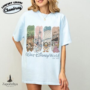 May include: Pale blue t-shirt with a graphic of Disney characters and landmarks. The shirt has the text "Walt Disney World" and "Est. 1971". The shirt is labelled "Comfort Colors Chambray".