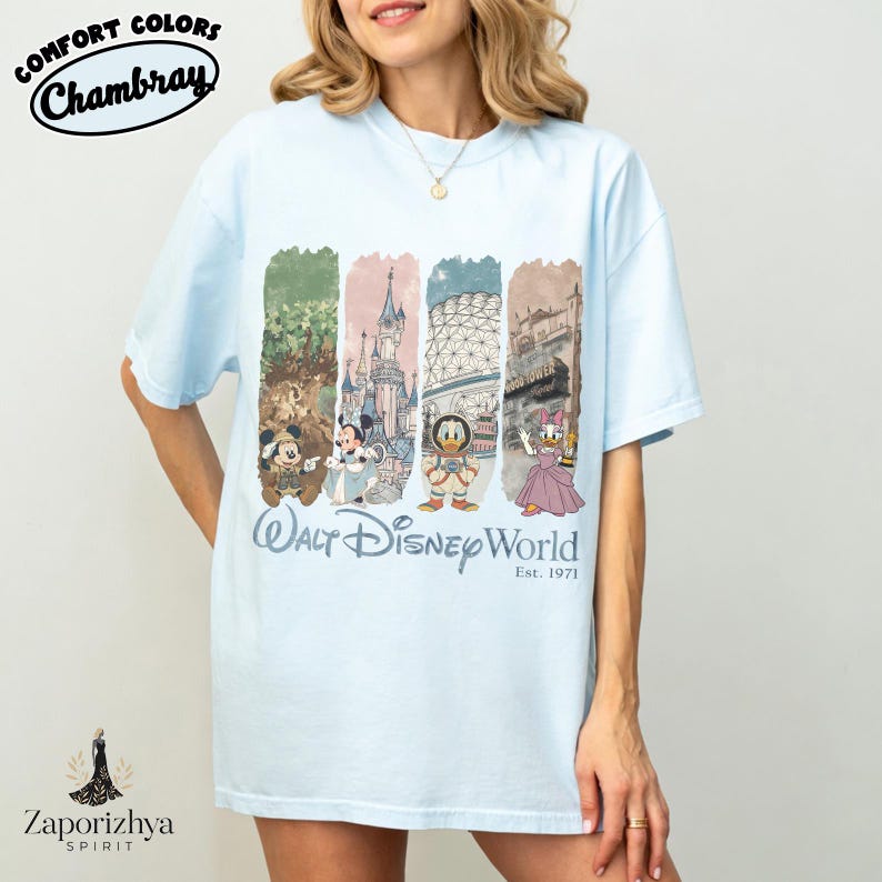 May include: Pale blue t-shirt with a graphic of Disney characters and landmarks. The shirt has the text "Walt Disney World" and "Est. 1971". The shirt is labelled "Comfort Colors Chambray".