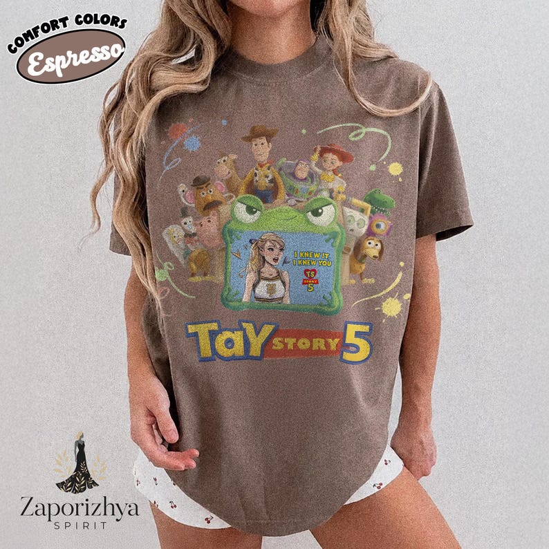 May include: A brown short-sleeve t-shirt featuring a graphic of various animated characters surrounding a green frog-shaped frame. Inside the frame is an illustration of a person with the text "I KNEW IT, I KNEW YOU" and "TAY STORY 5" below.