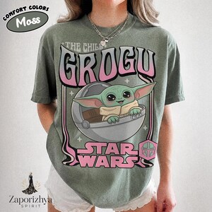 May include: A moss green short-sleeve t-shirt featuring a graphic print of Grogu in a floating pod. The text reads THE CHILD GROGU and STAR WARS in a pink and white gradient font. The design includes small star accents and a pink helmet icon.