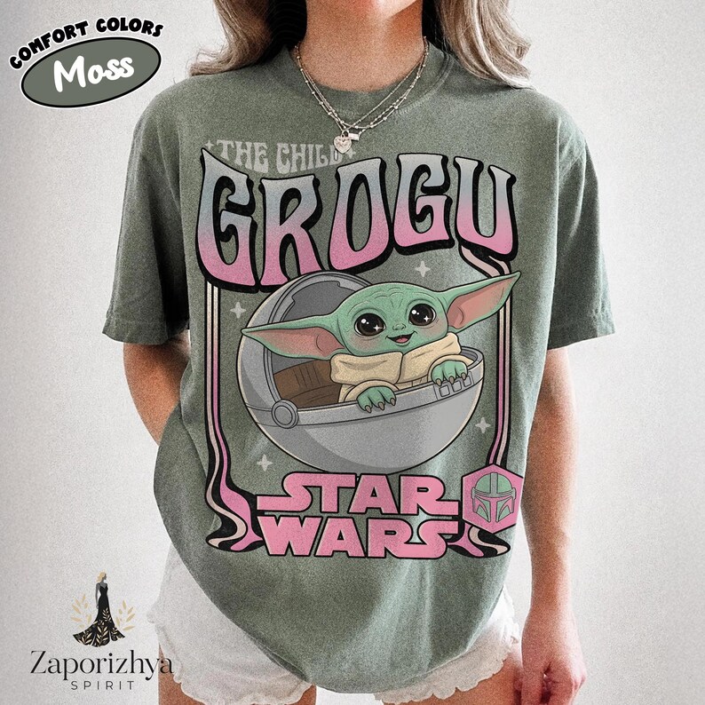 May include: A moss green short-sleeve t-shirt featuring a graphic print of Grogu in a floating pod. The text reads THE CHILD GROGU and STAR WARS in a pink and white gradient font. The design includes small star accents and a pink helmet icon.