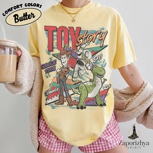 May include: A pale yellow t-shirt featuring a graphic print of Toy Story characters Woody, Buzz Lightyear, and Rex. The text reads Toy Story, Since 1995, Woody, Buzz, and Rex. The shirt is worn with a beige cardigan and pink checkered shorts.