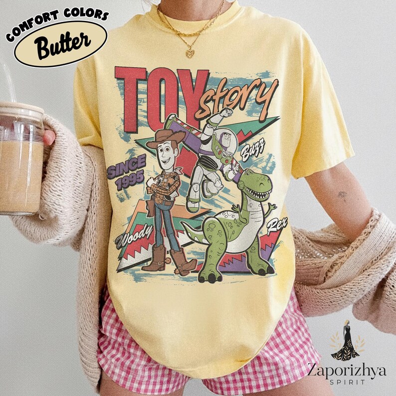 May include: A pale yellow t-shirt featuring a graphic print of Toy Story characters Woody, Buzz Lightyear, and Rex. The text reads Toy Story, Since 1995, Woody, Buzz, and Rex. The shirt is worn with a beige cardigan and pink checkered shorts.