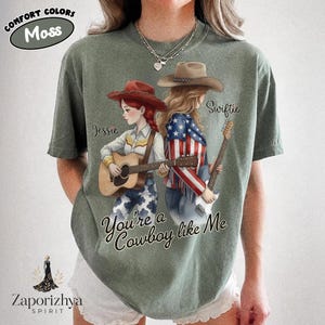 May include: A moss green t-shirt featuring a graphic of two characters with guitars. One character wears a red hat and cowgirl outfit, the other a brown hat and American flag jacket. Text reads "Jessie", "Swiftie", and "You're a Cowboy like Me".