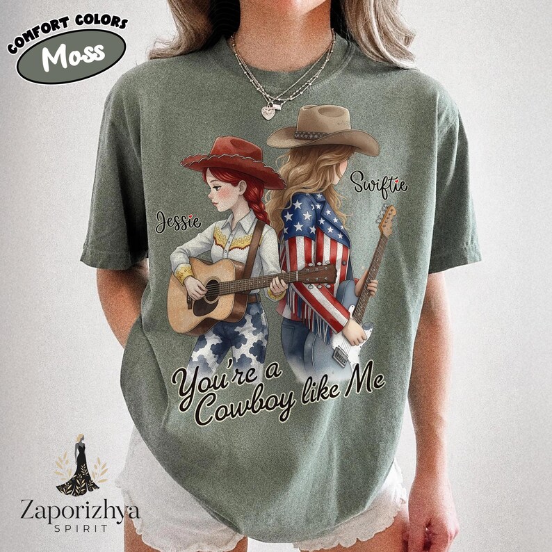 May include: A moss green t-shirt featuring a graphic of two characters with guitars. One character wears a red hat and cowgirl outfit, the other a brown hat and American flag jacket. Text reads "Jessie", "Swiftie", and "You're a Cowboy like Me".