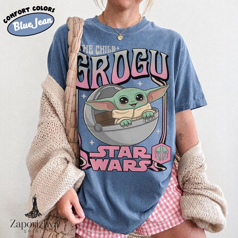 May include: A blue jean colored t-shirt featuring a graphic of Grogu from Star Wars inside a floating pod. The text reads THE CHILD, GROGU, and STAR WARS in pink and white lettering. The design includes small star accents and a helmet icon.