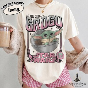 May include: An ivory t-shirt featuring a graphic of Grogu from Star Wars inside a floating pod. The text reads THE CHILD GROGU and STAR WARS in a pink and black gradient font. A small pink Mandalorian helmet icon is positioned near the bottom right.