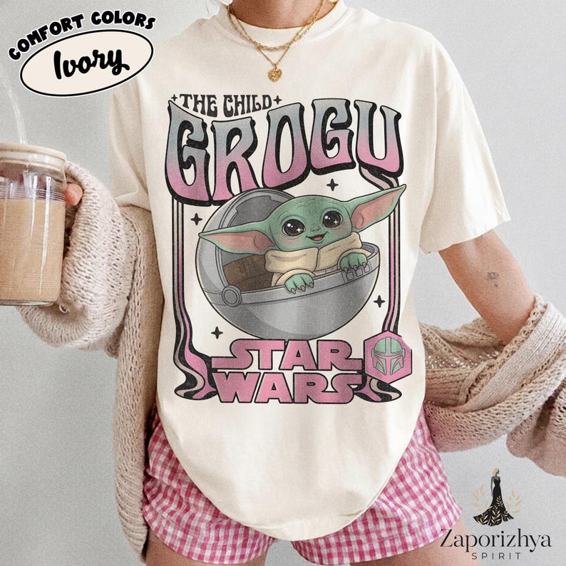 May include: An ivory t-shirt featuring a graphic of Grogu from Star Wars inside a floating pod. The text reads THE CHILD GROGU and STAR WARS in a pink and black gradient font. A small pink Mandalorian helmet icon is positioned near the bottom right.