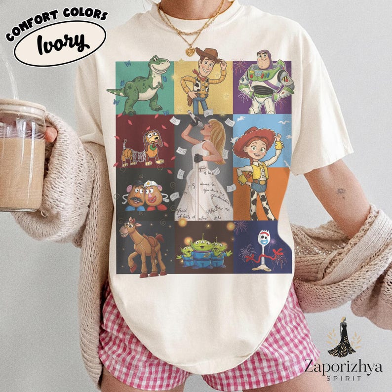 May include: An ivory short-sleeve t-shirt featuring a grid of nine colorful illustrations of animated characters, including a green dinosaur, a cowboy, a space ranger, a dog, a person in a white dress, a cowgirl, potato figures, a horse, aliens, and a spork.