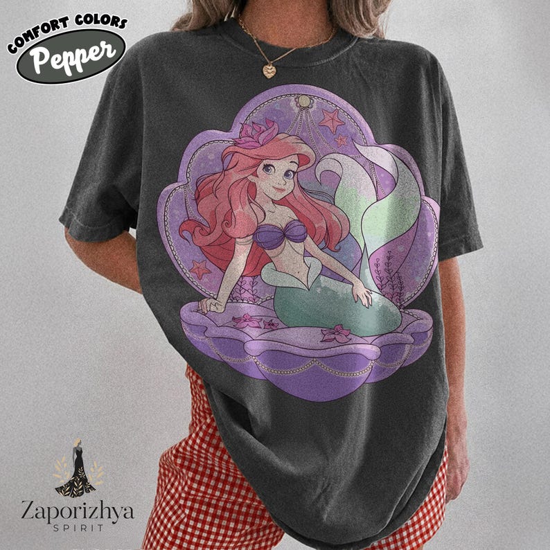 May include: A charcoal gray short-sleeve t-shirt featuring a graphic of a mermaid with long red hair, a purple seashell top, and a green tail, sitting inside a large purple seashell. The design includes small starfish and seaweed accents.