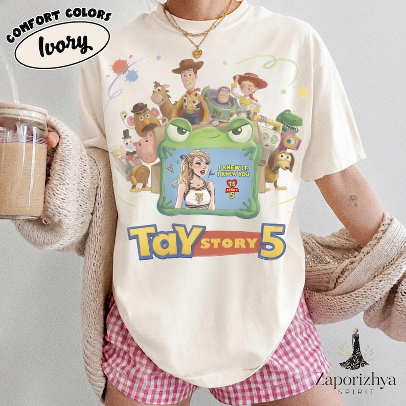 May include: An ivory t-shirt featuring a graphic print of various animated characters surrounding a green frog-shaped frame. Inside the frame is an illustration of a blonde person with the text I KNEW IT I KNEW YOU TS 5. Below is the text TAY STORY 5.