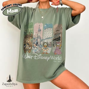 May include: A moss green t-shirt with a vibrant graphic showcasing Walt Disney World characters and landmarks. The design includes Mickey, Minnie, Donald, and Daisy Duck. The text "Walt Disney World Est. 1971" is printed below the graphic.