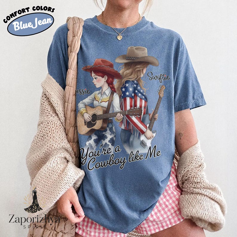 May include: A blue short-sleeve t-shirt featuring a graphic of two figures with guitars. One figure wears a red hat and the other a brown hat with a flag-patterned jacket. Text reads Jessie, Swiftie, and You're a Cowboy like Me.