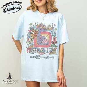 May include: Pale blue t-shirt with a colourful graphic of a purple dragon, the letter "D", and the words "Walt Disney World." The shirt also has the text "Comfort Colors Chambray" in a circular design.