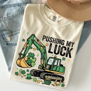May include: Cream-colored t-shirt with a green excavator graphic and the text "PUSHING MY LUCK". The excavator bucket is filled with shamrocks. Additional shamrocks, a pot of gold, and a horseshoe are also featured.