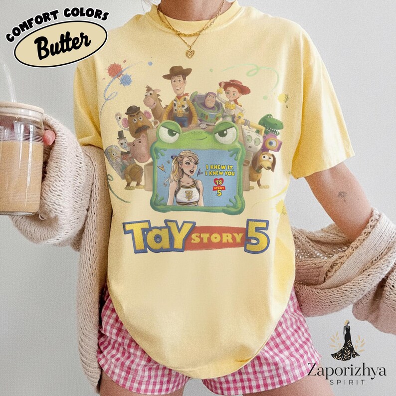May include: A butter yellow t-shirt featuring a graphic print of various animated characters surrounding a green frog frame. Inside the frame is a blonde person with the text "I KNEW IT, I KNEW YOU" and "TS 5". Below the graphic is the text "TAY STORY 5".
