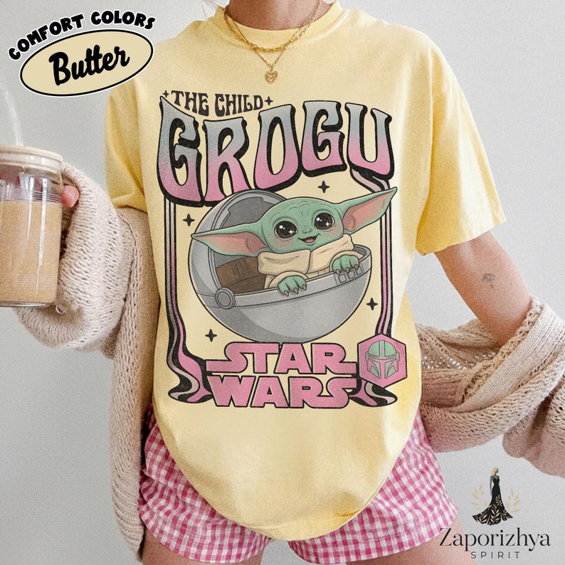 May include: A pale yellow t-shirt featuring a graphic of the character Grogu from Star Wars inside a floating pod. The text reads THE CHILD GROGU and STAR WARS in pink and black lettering. The design includes small star accents and a helmet icon.