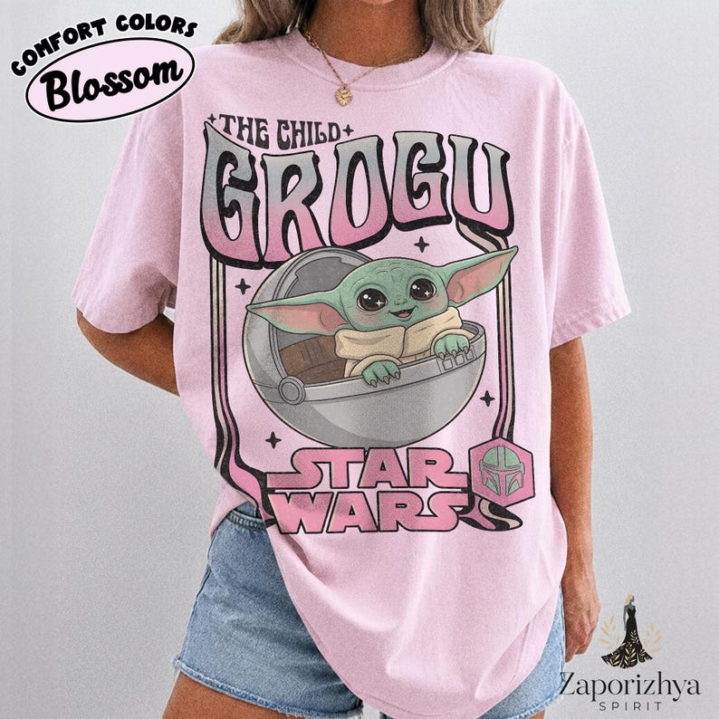 May include: A light pink short-sleeve t-shirt featuring a graphic of the character Grogu from Star Wars inside a floating pod. The text reads The Child, Grogu, and Star Wars, with a small helmet icon. The shirt has a crew neck and a relaxed fit.