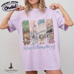 May include: A lilac t-shirt with a graphic of Disney characters and landmarks. The shirt features the text "Walt Disney World" and "Est. 1971". The top left corner has the text "Comfort Colors" and "Orchid".