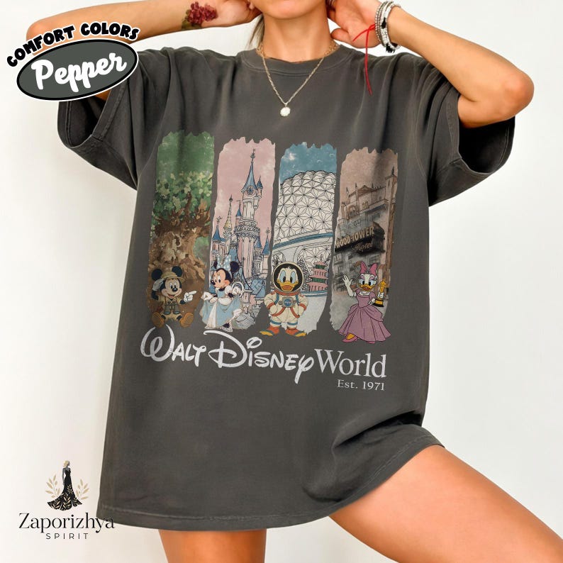 May include: Dark grey t-shirt with a colourful graphic featuring Disney characters and landmarks. The text "Walt Disney World Est. 1971" is printed below the graphic. The shirt has a relaxed fit and short sleeves.