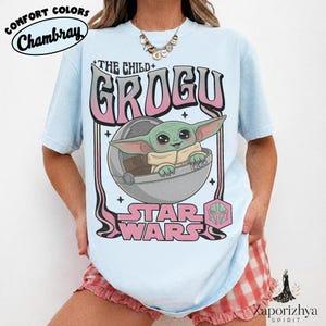May include: A light blue t-shirt featuring a graphic print of Grogu from Star Wars in a floating pod. The text reads THE CHILD, GROGU, and STAR WARS in pink and black lettering. The design includes a small pink hexagonal icon with a helmet.