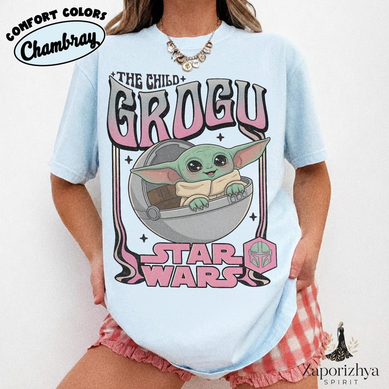 May include: A light blue t-shirt featuring a graphic print of Grogu from Star Wars in a floating pod. The text reads THE CHILD, GROGU, and STAR WARS in pink and black lettering. The design includes a small pink hexagonal icon with a helmet.