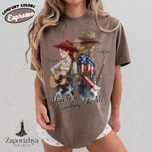 May include: A brown short-sleeve t-shirt featuring a graphic of two figures with cowboy hats playing acoustic guitars. One figure wears a red hat and the other a brown hat with a stars and stripes jacket. Text reads Jessie, Swiftie, and You're a Cowboy like Me.