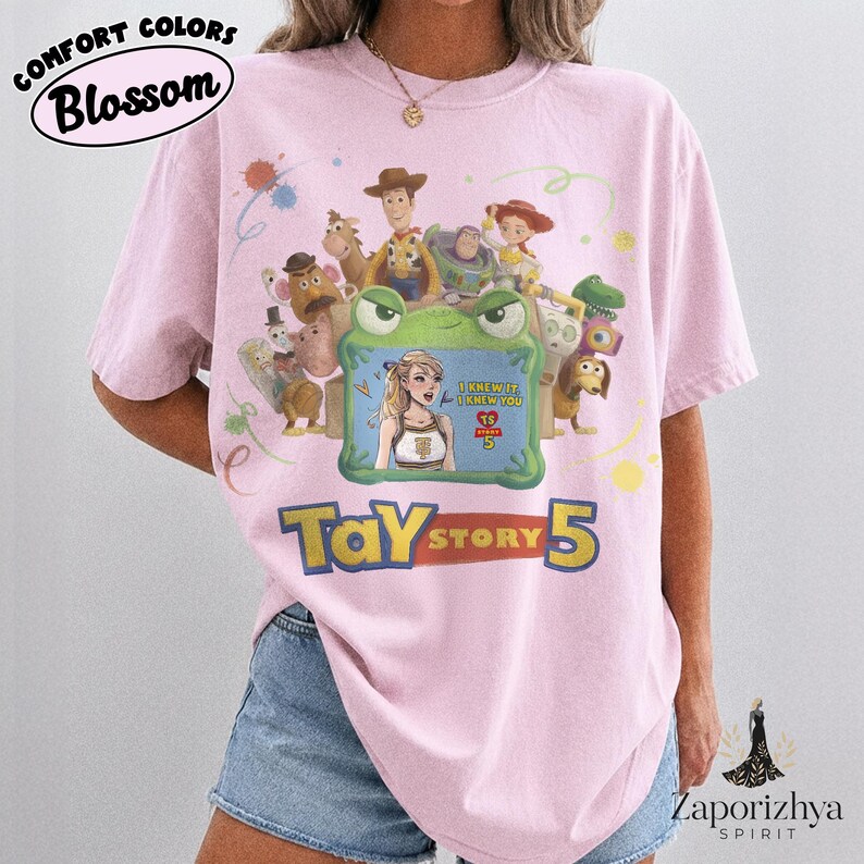 May include: A light pink t-shirt featuring a graphic print of various animated characters surrounding a green frog-shaped frame. Inside the frame is an illustration of a blonde person with the text I KNEW IT I KNEW YOU TS 5. Below the graphic is the text TAY STORY 5.