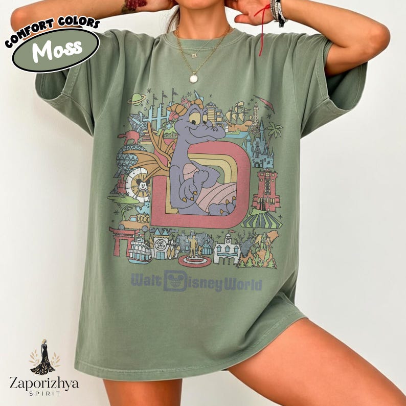 May include: A moss green t-shirt with a colourful graphic featuring a purple dragon, a rainbow, and various landmarks. The text "Walt Disney World" is printed below the graphic. The shirt has short sleeves and a relaxed fit.