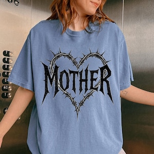May include: Pale blue t-shirt with a black barbed wire heart design and the word "MOTHER" in a gothic font. The shirt has a crew neck and short sleeves. The design is centered on the chest.
