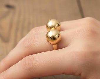 May include: A gold-toned ring featuring two smooth, polished spherical beads stacked vertically. The metal surface is reflective and glossy, creating a rounded, ball-like design that sits prominently on the finger. The band is simple and thin.