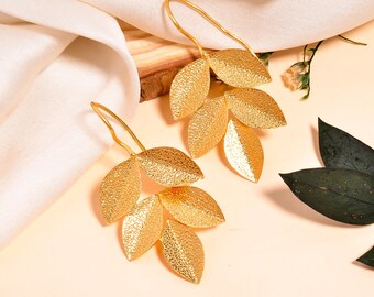 May include: A pair of gold-colored leaf earrings. Each earring features a cluster of textured, stylized leaves. The earrings have a long, curved hook for pierced ears. The leaves have a textured surface and a metallic sheen.