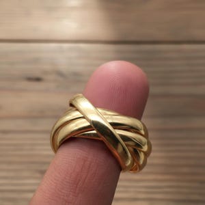 Può includere: Un anello in oro lucido caratterizzato da un design a più fasce con diversi anelli metallici lisci e intrecciati che si incrociano per creare un aspetto arrotondato e stratificato. L'anello presenta una finitura brillante e fluida.