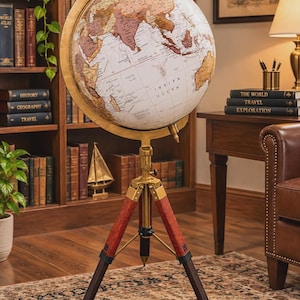 Puede incluir: Un globo terráqueo de pie con un mapa en tonos blanco y bronce, con etiquetas de texto negro para continentes y océanos. La esfera está sujeta por un anillo meridiano de latón y montada sobre un trípode de tres patas de madera oscura.