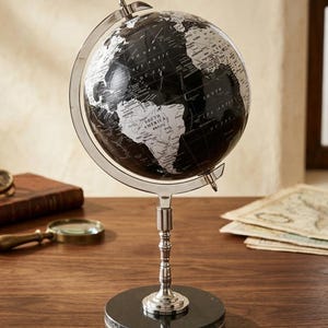 Op de afbeelding: Een zwart-witte wereldbol met zilverkleurige metalen accenten. De wereldbol is gemonteerd op een zilveren standaard met een zwarte marmeren voet. De continenten zijn wit op een zwarte achtergrond. Een decoratief item.